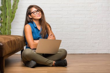 Kollarını, gülümseyen ve mutlu crossing, kendinden emin ve dostça katta oturan genç latin kadın portresi. Bir dizüstü bilgisayar holding.