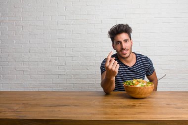 Pozitif ve güler yüzlü gel, kendinden emin ve gülümseyen bir hareketi el ile yapmak için davet bir masada oturan genç yakışıklı ve doğal bir adam. Taze bir salata yemek.