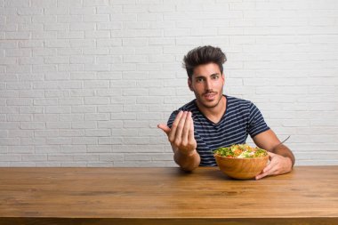Pozitif ve güler yüzlü gel, kendinden emin ve gülümseyen bir hareketi el ile yapmak için davet bir masada oturan genç yakışıklı ve doğal bir adam. Taze bir salata yemek.