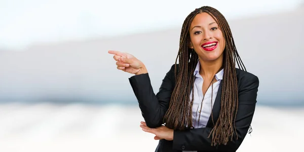 Portrait of a young black business woman pointing to the side, smiling surprised presenting something, natural and casual