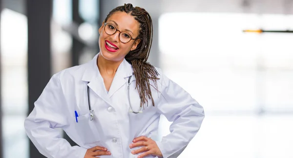 Portrait of a young black doctor woman with hands on hips, standing, relaxed and smiling, very positive and cheerful