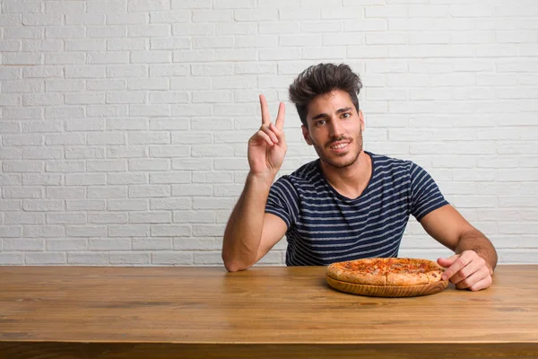 Young handsome and natural man sitting on a table fun and happy, positive and natural, doing a gesture of victory, peace concept. Eating a delicious pizza.