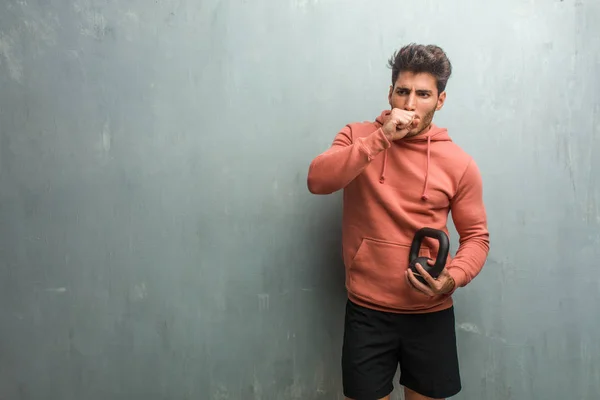 Young fitness man against a grunge wall with a sore throat, sick due to a virus, holding an iron dumbbell.