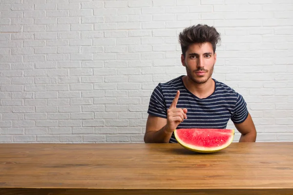 Young handsome and natural man sitting on a table showing number one, symbol of counting, concept of mathematics, confident and cheerful. Eating a watermelon.