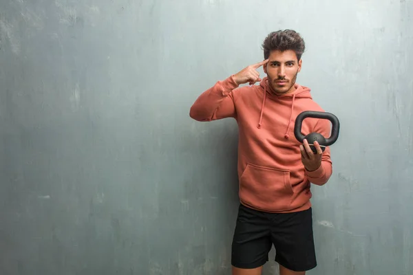 Young fitness man against a grunge wall man making a concentration gesture, holding an iron dumbbell.