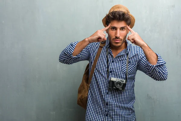Young handsome traveler man wearing a straw hat, a backpack and a photo camera man making a concentration gesture, looking straight ahead focused on a goal
