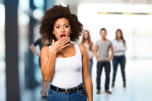 surprised young black woman covering her mouth with blurred people in background