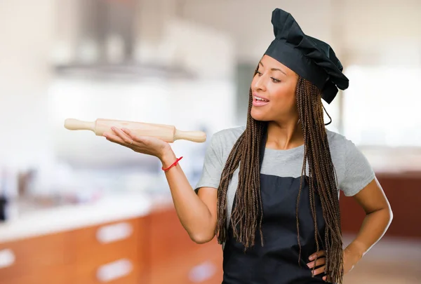 Portrait of a young black baker woman holding something with hands, showing a product, smiling and cheerful, offering an imaginary object