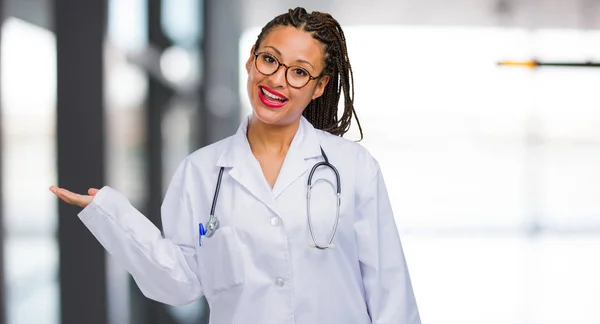 Portrait of a young black doctor woman holding something with hands, showing a product, smiling and cheerful, offering an imaginary object