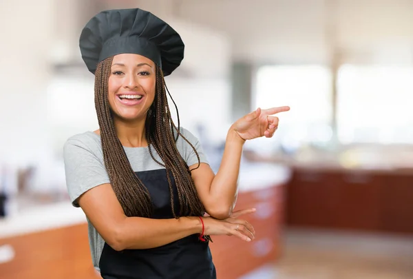 Portrait of a young black baker woman pointing to the side, smiling surprised presenting something, natural and casual