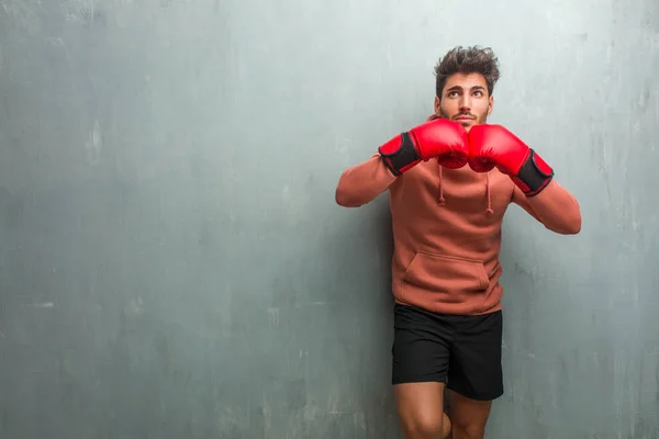 Young fitness man with boxing gloves against a grunge wall looking up, thinking of something