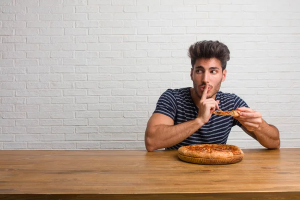 Young handsome and natural man sitting on a table keeping a secret or asking for silence, serious face, obedience concept. Eating a delicious pizza.