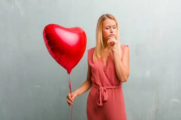 Portrait of young elegant blonde woman doubting and confused, thinking of an idea or worried about something. Holding a red heart balloon.