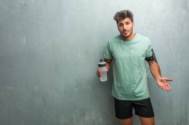 Telefon bir kolluk giyen genç fitness adam spor şişe şüphe ve omuzlar, shrugging bir grunge duvara.