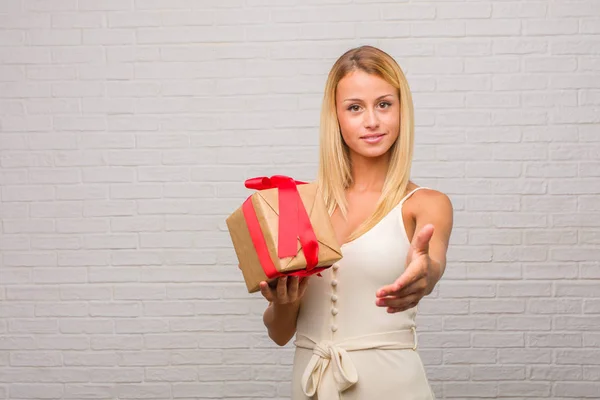 Portrait of young pretty blonde woman against a bricks wall reaching out to greet someone or gesturing to help, happy and excited. Holding a gift.