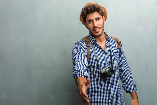 Young handsome traveler man wearing a straw hat, a backpack and a photo camera reaching out to greet someone or gesturing to help, happy and excited