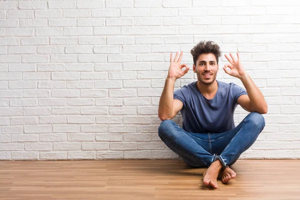 Young natural man sit on a wooden floor cheerful and confident doing ok gesture, excited and screaming, concept of approval and success