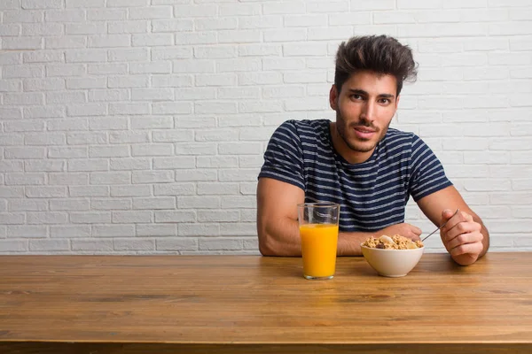 Kalça, rahat ve gülümseyen, çok olumlu ve neşeli ayakta, el ile bir masada oturan genç yakışıklı ve doğal adam. Bir kahvaltı yapıyor, portakal suyu ve tahıl bowl içerir.