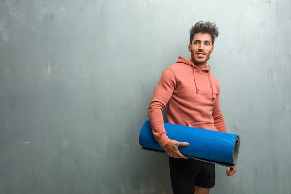 Young fitness man against a grunge wall crossing his arms, holding a blue mat for practicing yoga.