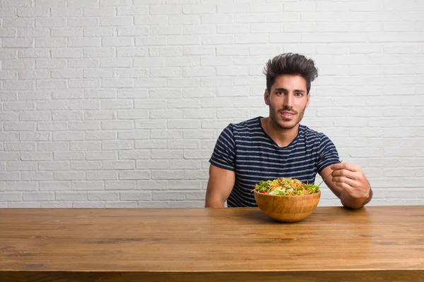 Young handsome and natural man sitting on a table with hands on hips, standing, relaxed and smiling, very positive and cheerful. Eating a fresh salad.