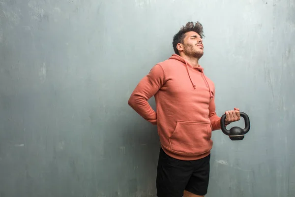 Young fitness man against a grunge wall with back pain due to work stress, tired and astute. Holding an iron dumbbell.