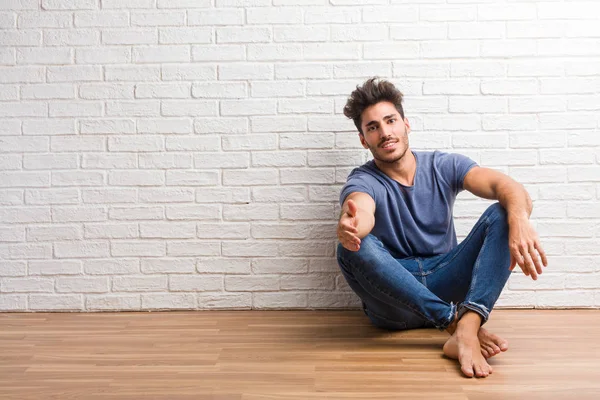 Young natural man sit on a wooden floor reaching out to greet someone or gesturing to help, happy and excited