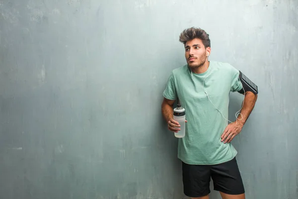 Young fitness man with sports bottle against a grunge wall doubting and shrugging shoulders, wearing an armband with phone.