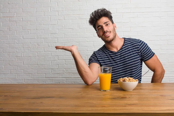 Young handsome and natural man sitting on a table holding something with hands, showing a product, smiling and cheerful, offering an imaginary object. Having a breakfast, includes orange juice and a cereals bowl.
