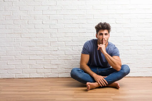 Young natural man sit on a wooden floor keeping a secret or asking for silence, serious face, obedience concept