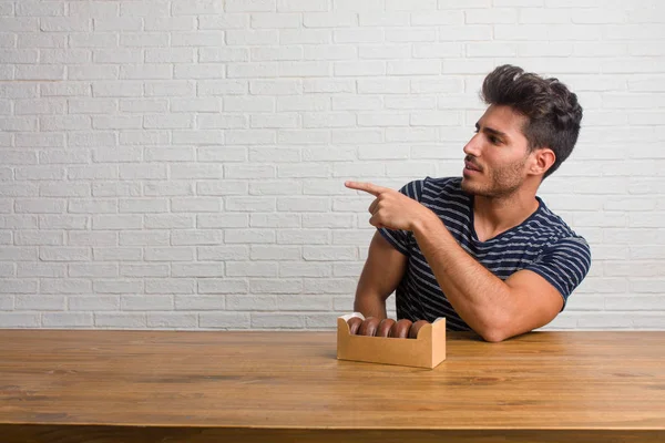 Young handsome and natural man sitting on a table pointing to the side, smiling surprised presenting something, natural and casual. Eating chocolate doughnuts.