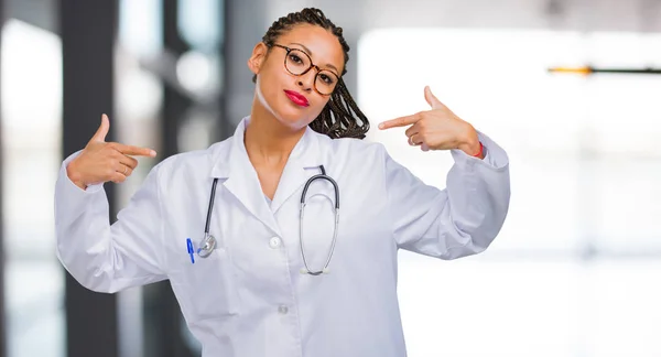 Portrait of a young black doctor woman proud and confident, pointing fingers, example to follow, concept of satisfaction, arrogance and health