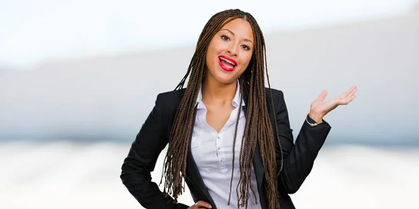 Portrait of a young black business woman holding something with hands, showing a product, smiling and cheerful, offering an imaginary object