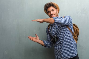 Hasır şapka, bir sırt çantası ve el ile bir şey tutan bir fotoğraf makinesi giyen genç yakışıklı gezgin adam bir ürün göstermek gülümseyen ve neşeli, hayali bir nesne sunan