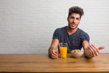 Pozitif ve güler yüzlü gel, kendinden emin ve gülümseyen bir hareketi el ile yapmak için davet bir masada oturan genç yakışıklı ve doğal bir adam. Bir kahvaltı yapıyor, portakal suyu ve tahıl bowl içerir.