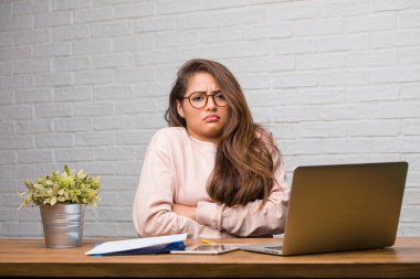 Şüphe ve omuzlarında shrugging masada oturan genç öğrenci latin kadın portresi