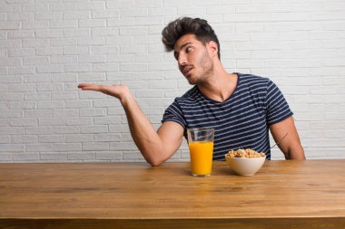 Elleriyle bir şey tutan, bir ürün, gülümseyen ve neşeli gösterilen, hayali bir nesne sunan bir masada oturan genç yakışıklı ve doğal adam. Bir kahvaltı yapıyor, portakal suyu ve tahıl bowl içerir.