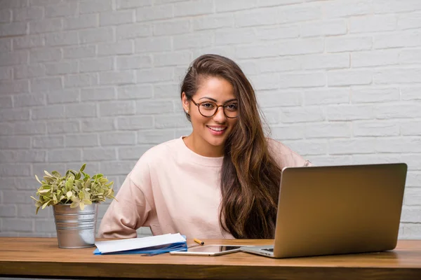 Portrait of young student latin woman sitting on her desk with hands on hips, standing, relaxed and smiling, very positive and cheerful