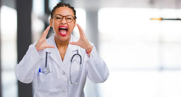 Portrait of a young black doctor woman screaming angry, expression of madness and mental instability, open mouth and half-opened eyes, madness concept