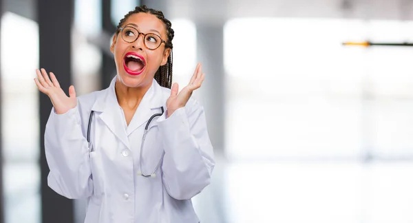 Portrait of a young black doctor woman surprised and shocked, looking with wide eyes, excited by an offer or by a new job, win concept