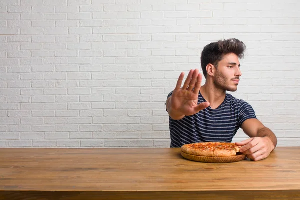 Young handsome and natural man sitting on a table serious and determined, putting hand in front, stop gesture, deny concept. Eating a delicious pizza.