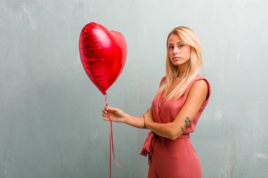 Kendinden emin ve göstermek güç hissi ve heybetli, kollarını, ciddi geçiş zarif sarışın genç kadın portresi. Bir kırmızı kalp balon holding.