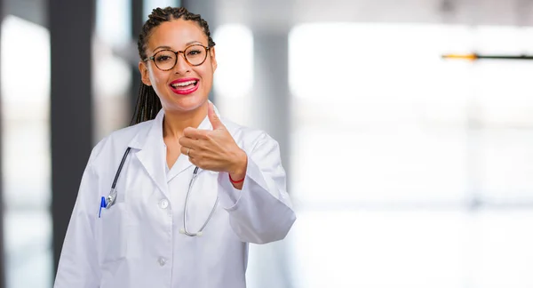 Portrait of a young black doctor woman cheerful and excited, smiling and raising her thumb up, concept of success and approval, ok gesture