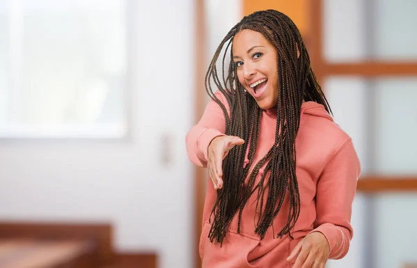 Portrait of a young black woman wearing braids reaching out to greet someone or gesturing to help, happy and excited