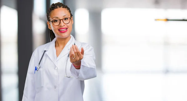 Portrait of a young black doctor woman inviting to come, confident and smiling making a gesture with hand, being positive and friendly