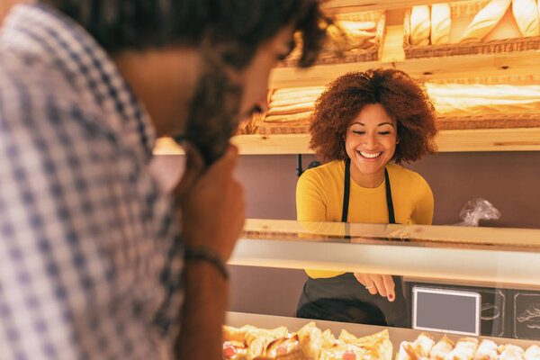 Young man buying something in a bakery, is being attended by an pretty afro woman.