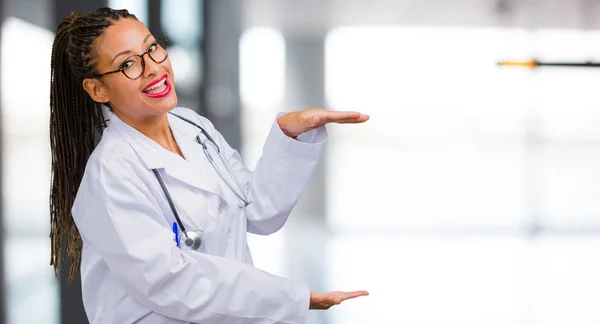 Portrait of a young black doctor woman holding something with hands, showing a product, smiling and cheerful, offering an imaginary object