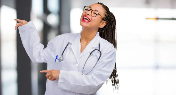 Portrait of a young black doctor woman pointing to the side, smiling surprised presenting something, natural and casual
