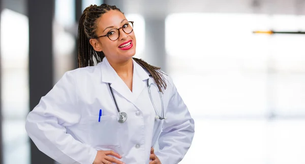 Portrait of a young black doctor woman with hands on hips, standing, relaxed and smiling, very positive and cheerful