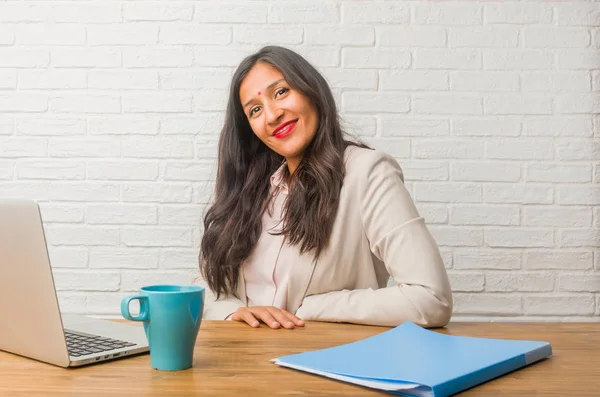 Young indian woman at the office with hands on hips, standing, relaxed and smiling, very positive and cheerful