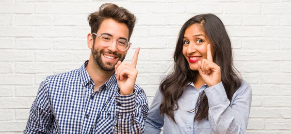 Young indian woman and caucasian man couple showing number one, symbol of counting, concept of mathematics, confident and cheerful